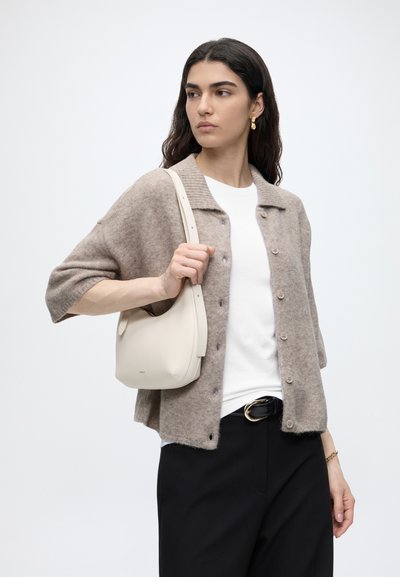 Femme aux cheveux foncés portant un cardigan beige boutonné, une chemise blanche, un pantalon noir et des boucles d'oreilles dorées, tenant un petit sac à épaule crème.