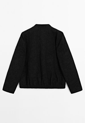 Veste noire à manches longues avec un motif floral subtil, ourlet élastique et sans fermetures visibles, vue de dos sur fond blanc.