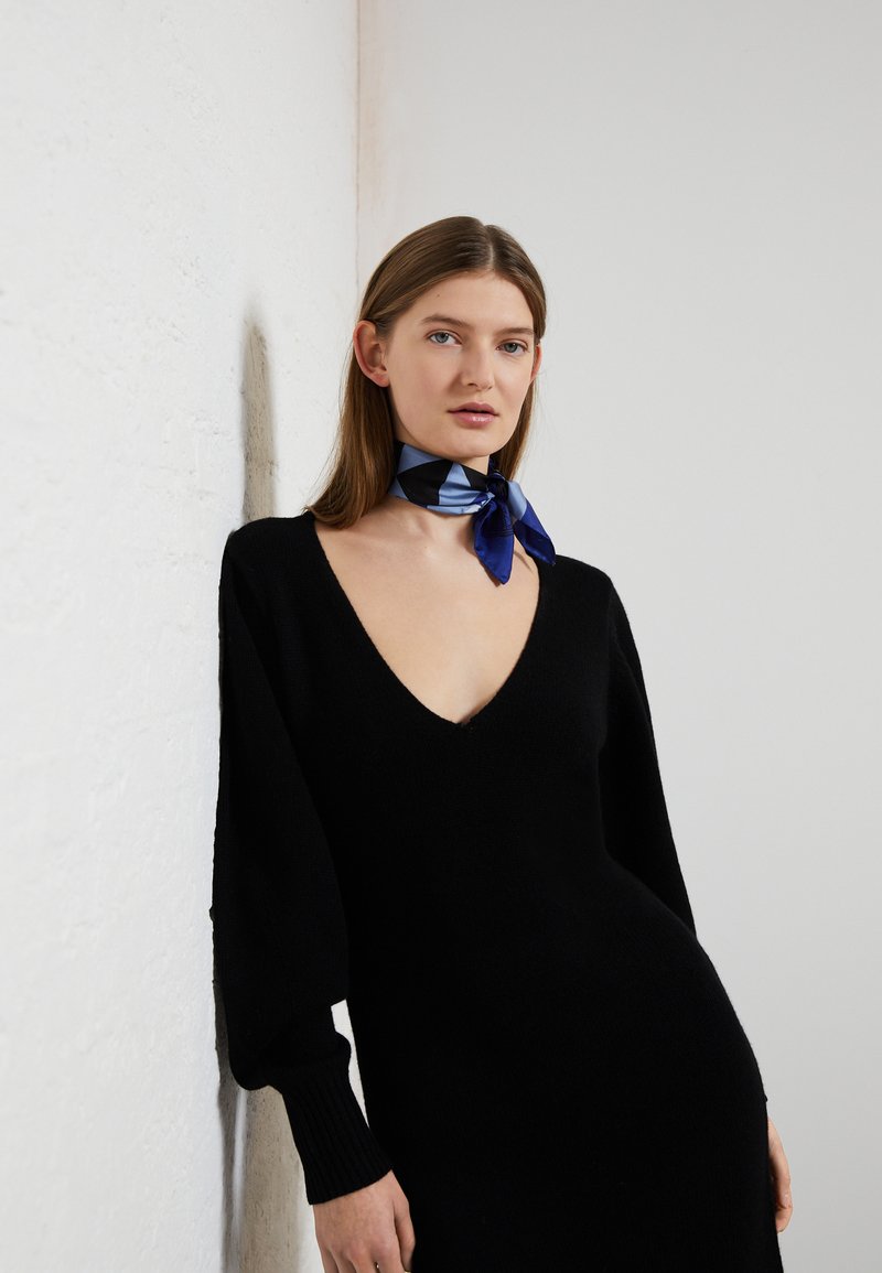 Vestido preto de malha com um decote em V profundo e mangas bufantes. Combinado com um lenço de seda azul com padrões geométricos amarrado ao redor do pescoço.