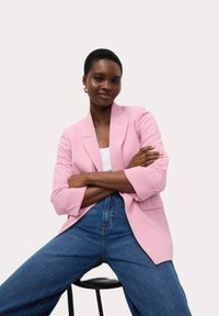 Blazer ajusté rose clair avec revers, plis aux manches, associé à un jean taille haute bleu, sur un tabouret noir contre un fond neutre.