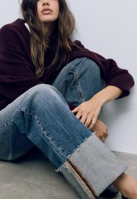 Mörkröd tröja, löst sittande ljusblå jeans med fransade kanter och kontrasterande orange rand längs fållarna, bärs med svarta skor.