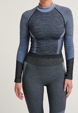 Person wearing form-fitting, long-sleeve blue-gray athletic top and matching high-waisted leggings, standing against plain background.