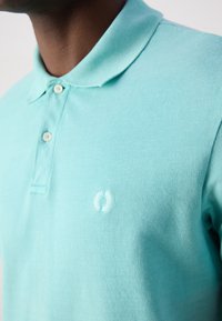 Polo turquoise en tissu texturé, avec un col doux, trois boutons et un logo brodé discret sur la poitrine.