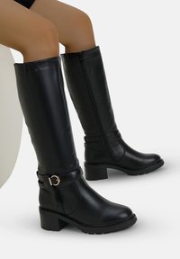 Botas negras altas hasta la rodilla hechas de cuero sintético suave, con punta redonda, tacón cuadrado y cremallera lateral. Incluyen un acento decorativo en forma de correa.