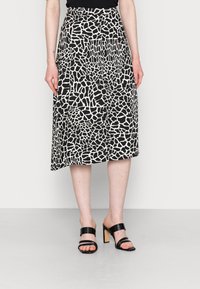 Schwarz-weißer Midi-Rock mit giraffenähnlichem Muster, fließendem Design und hoher Taille, kombiniert mit schwarzen Absatzsandalen.