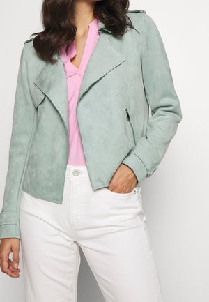 Mujer que lleva una chaqueta de ante verde claro sobre una blusa rosa y pantalones blancos, con una mano en el bolsillo.