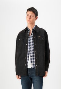 Črna denim jakna s srebrnimi snaps gumbi, z dvema žepoma na prsih, oblečena čez modro-belo karirano srajco in belo majico.
