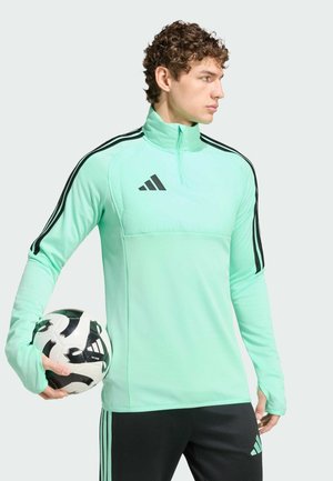 Muž v mentolově zeleném sportovním pulovru s černými pruhy drží černo-bílý fotbalový míč, tváří doprava na jednobarevném pozadí.