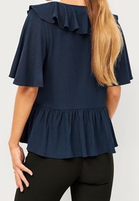 Kvinna iförd en marinblå texturerad blus med volangkrage och nederkant, kombinerad med svarta tighta byxor, sedd bakifrån.