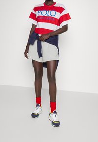 Person klädd i en randig Polo Ralph Lauren-t-shirt i röd och vit, grå shorts, röda strumpor, multifärgade sneakers och en marinblå sweatshirt knuten runt midjan.