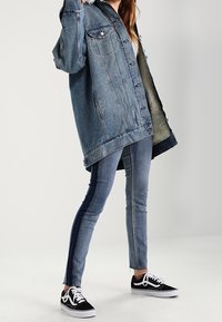 Nadměrná modrá džínová bunda s podšívkou z umělé ovčiny, kombinovaná s úzkými dvoubarevnými džínami a černými teniskami s bílými doplňky.