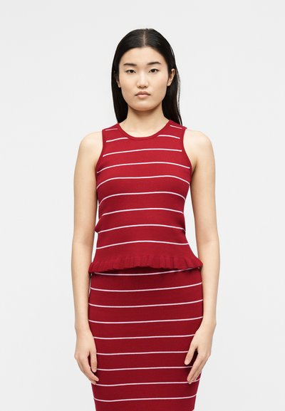 Νεαρή γυναίκα με κόκκινο αμάνικο τοπ και ταιριαστή φούστα με λευκές οριζόντιες ρίγες, στέκεται μπροστά σε απλό λευκό φόντο.