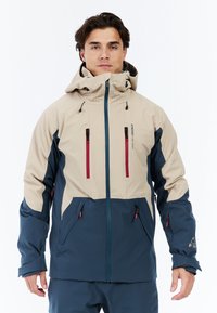 Beige en marineblauwe waterdichte jas met capuchon, met ritszakken en verstelbare manchetten. Heeft contrasterende panelen en ventilatie bij de kraag.