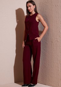 Vesta fără mâneci în nuanță de burgund, cu nasturi în față, asortată cu pantaloni evazați. Materialul neted și cele două buzunare laterale îmbunătățesc designul.