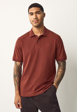 Mann in rostfarbenem Poloshirt mit tätowierten Armen, steht vor schlichtem hellem Hintergrund mit einer Hand in der Tasche.