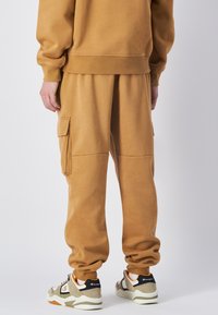 Braune Baumwoll-Sweatpants mit lockerem Schnitt, ausgestattet mit seitlichen Cargo-Taschen, elastischen Bündchen und kombiniert mit beigen und schwarzen Sneakers.