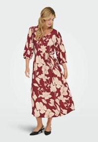 Maxiklänning i vinröd med krämfärgad blommönster, med V-ringning, 3/4 puffärmar och en cinchad midja för form. Svarta klackar fulländar looken.