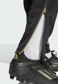 Zwarte sportbroek met een ritssluiting bij de enkel, die een wit gestreepte sok en een zwarte voetbalschoen met textuuraccenten en branding onthult.