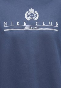 Μπλε ύφασμα με λευκό κεντημένο κείμενο "NIKE CLUB SINCE 1972" και έμβλημα με κλάδο δάφνης, στέμμα και λογότυπο Nike swoosh.