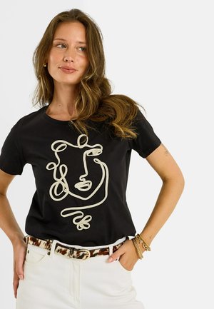 Femme aux longs cheveux ondulés portant un t-shirt noir avec un dessin abstrait de visage en ligne, un pantalon blanc, une ceinture imprimé léopard et des bracelets dorés.