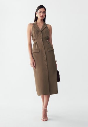 Vrouw met stijl donker haar die een mouwloze bruine midijurk met reverskraag en knopen draagt, een zwarte handtas vasthoudt en beige sandalen met hoge hakken draagt.