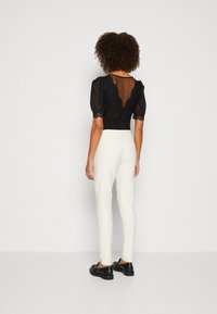 Blouse noire transparente avec manches bouffantes, détails en dentelle et motif à pois, associée à un pantalon blanc ajusté et des chaussures noires.