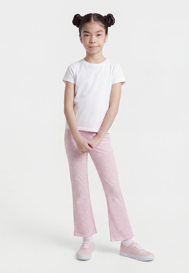 Νεαρή κοπέλα που στέκεται με σταυρωμένα τα χέρια, φοράει λευκό μπλουζάκι, ροζ παντελόνια με σχέδια και ροζ αθλητικά παπούτσια, μπροστά σε απλό λευκό φόντο.