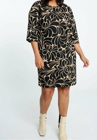 Femme portant une robe beige à mi-mollet avec un audacieux motif floral abstrait noir et des bottines beiges avec des talons noirs.