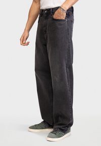 Jean en denim noir à coupe décontractée, design à cinq poches, coutures visibles et détail de marque subtil sur la cuisse. Associé à des baskets vertes.