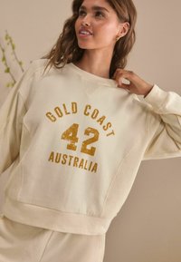 Krämfärgad sweatshirt med en texturerad design, rund halsringning och guldtryckt text som säger "GOLD COAST 42 AUSTRALIEN."