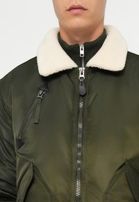 Veste bomber vert olive avec un col en fausse fourrure crème, fermeture éclair double et détails de poches zippées texturées sur le côté gauche.