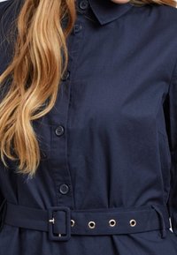 Marinblå skjorta med knappar och krage, med bälte i midjan och guldfärgade öglor. Slätt tyg med klassisk design.