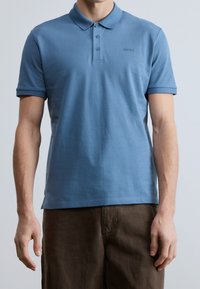 Homme portant un polo bleu à manches courtes avec trois boutons et un pantalon marron foncé, debout devant un fond uni.