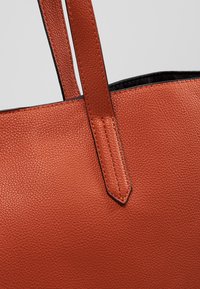 Gros plan sur un sac fourre-tout en cuir orange brûlé texturé avec des poignées cousues et une bordure noire le long du bord supérieur.