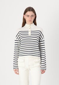 Strikket genser i kremfarge med marineblå striper, med høyt krage og fire gullknapper ved halsen. Myk tekstur, kort passform.