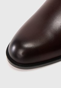 Pohled zblízka na špičku hladké tmavě hnědé kožené boty zaobleného tvaru s černým vroubkovaným okrajem podrážky na bílém pozadí.