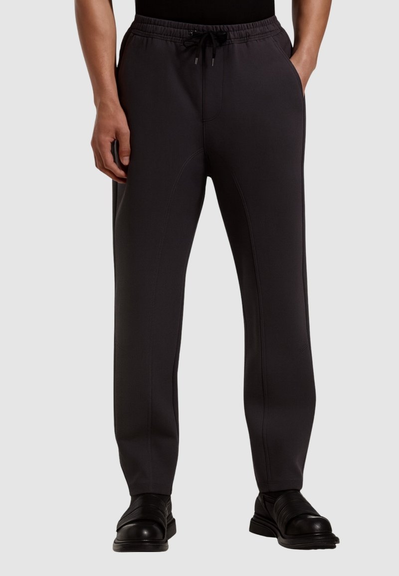 Pantaloni jogger grigio scuro con elastico in vita e cordoncino, dotati di cuciture laterali e vestibilità affusolata, abbinati a scarpe nere.