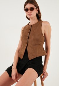 Barna velúr mellény kerek nyakkivágással, gombos elülső résszel és két oldalsó zsebbel, fekete rövidnadrággal párosítva. A modellen kerek napszemüveg van.