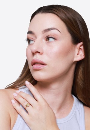 Frau mit langen braunen Haaren schaut seitlich, trägt ein weißes Tanktop und einen goldenen herzförmigen Ring an der linken Hand, die auf ihrer Schulter ruht.