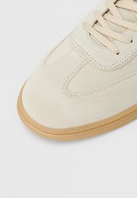 Šviesiai smėlio spalvos zomšinė sportinė avalynė su smėlio spalvos raišteliais ir guminiais padais, rodantis detalias siūles ir tekstūruotą pado kraštą.