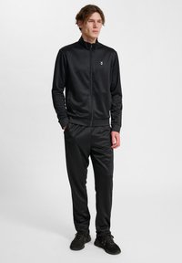 Svart atletisk träningsdräkt i ett slätt tyg. Jackan har en hög krage och dragkedja, byxorna har ett elastiskt midjeband. Enkelt design.