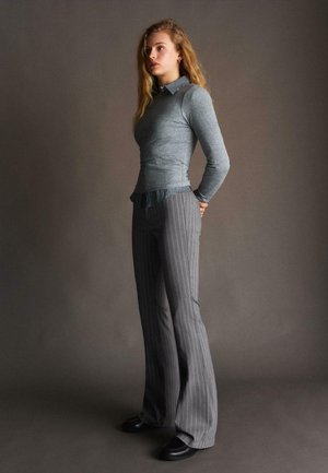 Giovane donna con lunghi capelli biondi, in piedi davanti a uno sfondo semplice, indossa un maglione grigio, una camicia a righe, pantaloni svasati a righe sottili e scarpe nere.