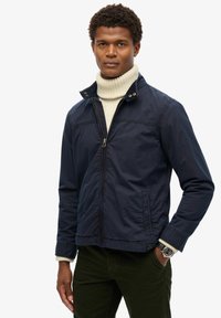 Mørkeblå zip-up jakke med ståkrave, fremstillet af et glat stof. Har to sidelommer og ribbede manchetter.