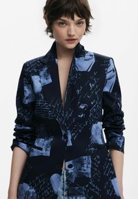 Blazer bleu marine avec un mélange de motifs bleus et de texte écrit à la main. Présente une coupe ajustée, des revers crantés et des manches retroussées, fabriqué en tissu lisse.