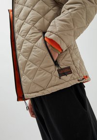 Doudoune matelassée beige avec doublure orange, poches zippées et étiquette logo. Présente une texture légèrement brillante et des poignets élastiques.