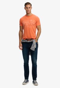 Camiseta naranja de cuello redondo con texto impreso en blanco, jeans de mezclilla oscuros y una camisa de cuadros atada a la cintura. Zapatillas casuales completan el conjunto.