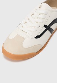 White leather sneaker with beige suede toe cap, black side stripes, and white laces. Rubber sole with a gum-colored bottom.
