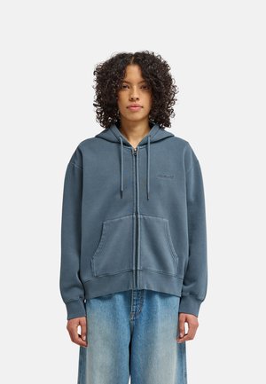 Persona con capelli ricci indossa una felpa blu con zip, coulisse e tasche frontali, abbinata a jeans blu larghi, in piedi davanti a uno sfondo semplice.
