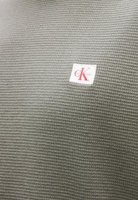 Γκρι υφασμάτινη υφή με ένα μικρό λευκό σημείο που φέρει κόκκινα γράμματα "cK" και το κείμενο "Calvin Klein" σε ανοιχτό γκρι χρώμα.