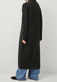 Černý dlouhý cardigan s texturovaným pleteným vzorem, dlouhými rukávy a rovného střihu, doplněný širokými modrými džíny.